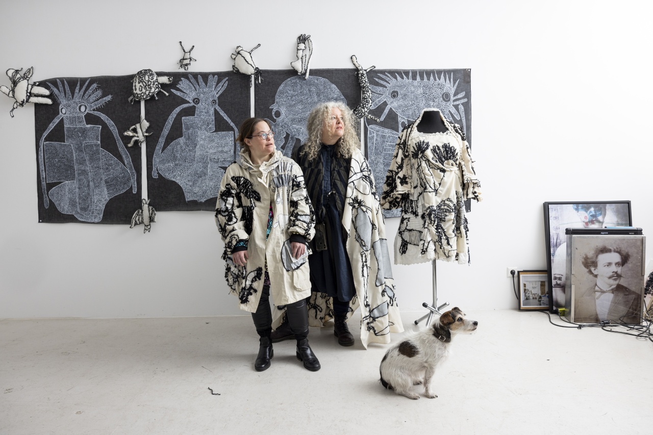2 Frauen in bestickter Kleidung stehen nebeneinander in einem Kunstatelier. Auf dem Boden sitzt ein Hund.